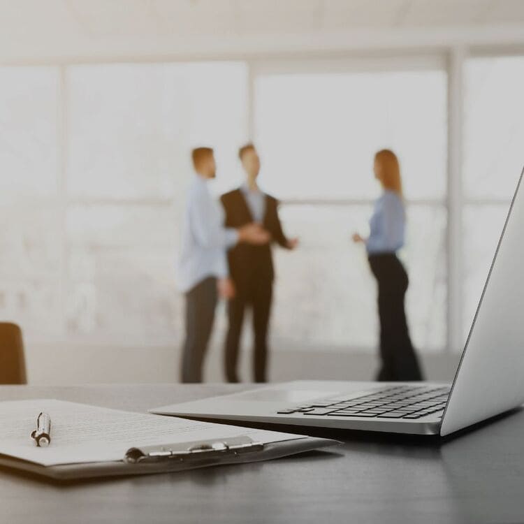 Ein Laptop und Notizen stehen im Vordergrund, während in der Hintergrund eine Gruppe von Mitarbeitern der CodeKlar GmbH diskutiert.